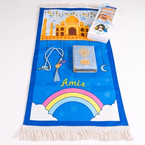 Peut inclure: Un tapis de pri&egrave;re bleu avec un arc-en-ciel et une mosqu&eacute;e, un chapelet et un petit livre. Le tapis porte le nom "Amir". Une bo&icirc;te avec le texte "Eid Mubarak" est &eacute;galement pr&eacute;sente.