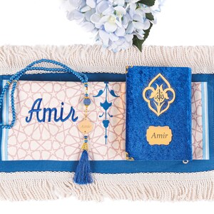 May include: A blue prayer mat with a cream-colored fringe, featuring the name "Amir" embroidered in blue. A blue beaded tasbih and a blue velvet Quran with gold accents are also present. A blue tassel and a floral arrangement complete the scene.