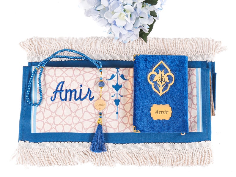 May include: A blue prayer mat with a cream-colored fringe, featuring the name "Amir" embroidered in blue. A blue beaded tasbih and a blue velvet Quran with gold accents are also present. A blue tassel and a floral arrangement complete the scene.