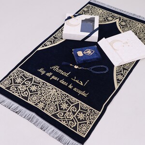 May include: A blue prayer rug with gold embroidery and a tassel. The rug has the name "Ahmed" and the Arabic phrase "May all your duas be accepted" embroidered on it. The rug is accompanied by a blue prayer book, a set of prayer beads, and a white gift box.