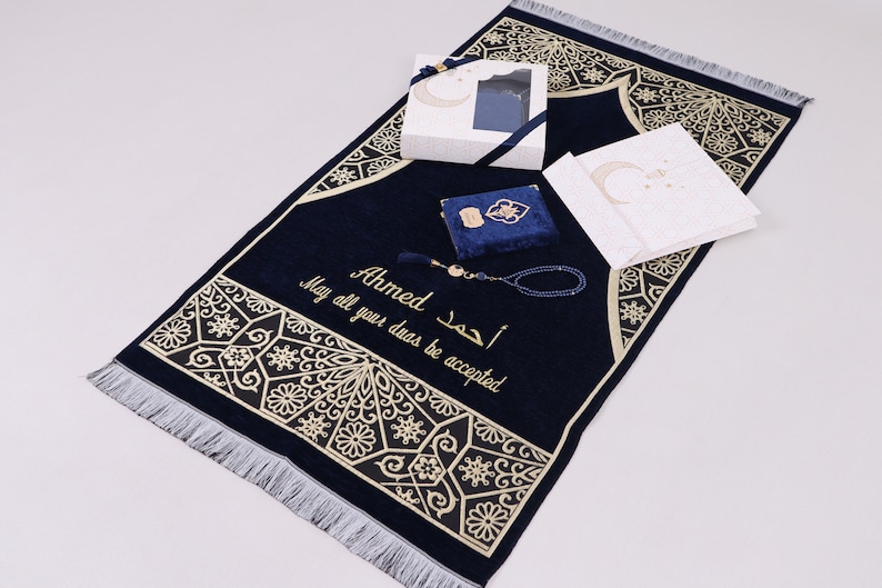 May include: A blue prayer rug with gold embroidery and a tassel. The rug has the name "Ahmed" and the Arabic phrase "May all your duas be accepted" embroidered on it. The rug is accompanied by a blue prayer book, a set of prayer beads, and a white gift box.