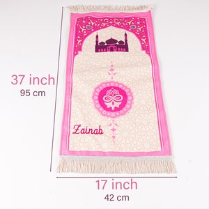 Peut inclure: Tapis de pri&egrave;re rose, de 95 cm de long, avec un motif de mosqu&eacute;e et le nom "Zainah". Le tapis a un fond cr&egrave;me avec un motif g&eacute;om&eacute;trique, des bordures roses et des franges. Il mesure 42 cm de large.