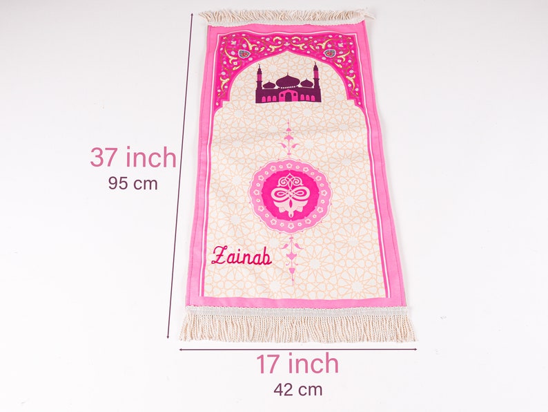 Peut inclure: Tapis de pri&egrave;re rose, de 95 cm de long, avec un motif de mosqu&eacute;e et le nom "Zainah". Le tapis a un fond cr&egrave;me avec un motif g&eacute;om&eacute;trique, des bordures roses et des franges. Il mesure 42 cm de large.