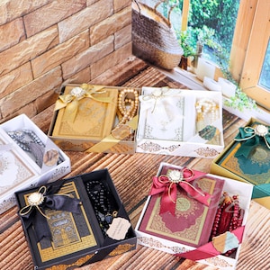 K&ouml;nnte beinhalten: Sechs Geschenkboxen mit einem Koran und Gebetsperlen. Die Boxen sind mit B&auml;ndern und Schleifen verziert. Die Boxen sind in verschiedenen Farben, darunter Gold, Wei&szlig;, Schwarz, Rot und Gr&uuml;n.