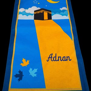 May include: A blue prayer rug with a yellow and orange design featuring the Kaaba, a crescent moon, and stars. The name "Adnan" is written in blue script. The rug has a fringed edge and maple leaf designs.