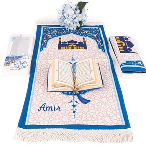 May include: A prayer rug with a mosque design, a Quran, and a prayer bead. The rug is beige with a blue border and a mosque illustration. The Quran is open, and the prayer beads are draped over it. A gift box with text is on the left.