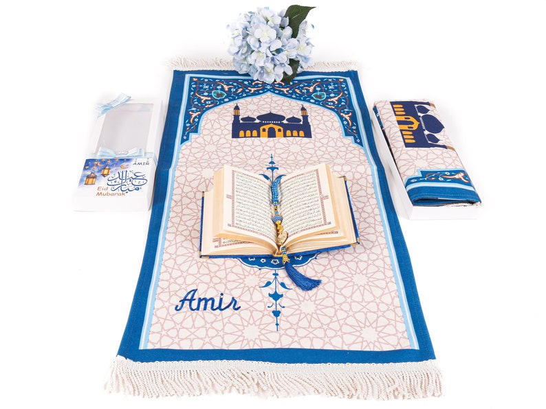 May include: A prayer rug with a mosque design, a Quran, and a prayer bead. The rug is beige with a blue border and a mosque illustration. The Quran is open, and the prayer beads are draped over it. A gift box with text is on the left.