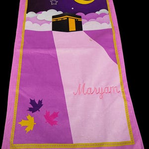 May include: A rectangular prayer rug with a purple and pink design. The top features a night sky with a crescent moon, stars, and clouds above a black building with a gold door. The name "Maryam" is embroidered in pink.