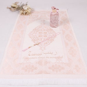 May include: A pink prayer rug with a white fringe, a pink prayer bead necklace, and a pink pouch with a white lace design. The rug has a gold-coloured floral design and the text "La ilaha illa Allah" in Arabic script, which translates to "There is no god but God".