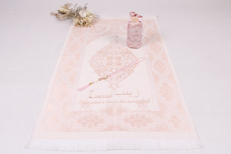 May include: A pink prayer rug with a white fringe, a pink prayer bead necklace, and a pink pouch with a white lace design. The rug has a gold-coloured floral design and the text "La ilaha illa Allah" in Arabic script, which translates to "There is no god but God".