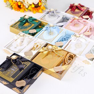 May include: Assortment of gift boxes with Quran books and prayer beads. The boxes are decorated with ribbons, bows, and gold accents. The books are in various colors, including green, blue, white, and gold. The prayer beads are white, black, and brown.