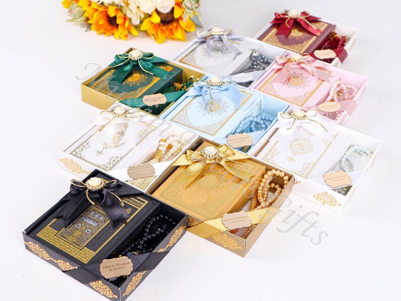 May include: Assortment of gift boxes with Quran books and prayer beads. The boxes are decorated with ribbons, bows, and gold accents. The books are in various colors, including green, blue, white, and gold. The prayer beads are white, black, and brown.