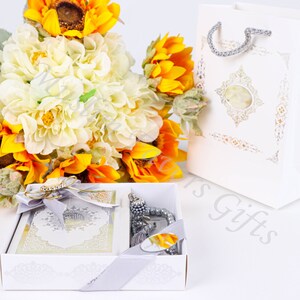 May include: A white gift box with a silver ribbon and a white gift bag with a gold design. The box contains a white prayer book with a silver design and a silver prayer beads. The box is surrounded by a bouquet of white and yellow flowers.