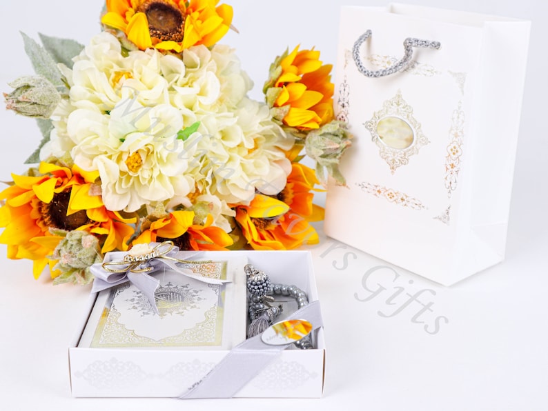 May include: A white gift box with a silver ribbon and a white gift bag with a gold design. The box contains a white prayer book with a silver design and a silver prayer beads. The box is surrounded by a bouquet of white and yellow flowers.