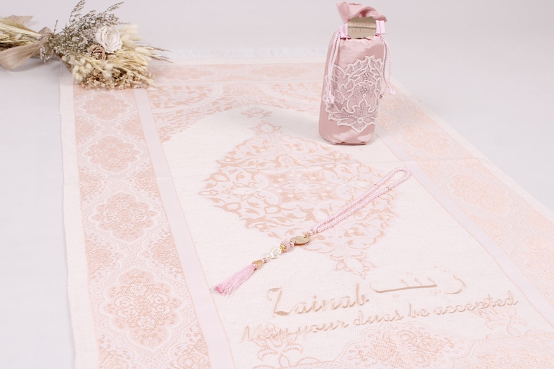 May include: A pink prayer rug with a delicate floral pattern and a matching pink prayer bag. The rug has the words "Lailaha" and "May your deeds be accepted" written on it. A pink prayer bead necklace with a tassel is on the rug.