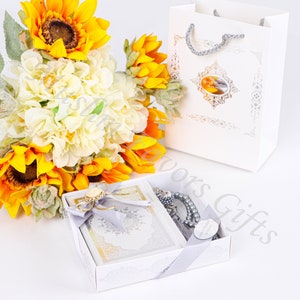 May include: A white gift box with a silver ribbon and a white gift bag. The box contains a small book and a silver prayer beads. A bouquet of sunflowers and white flowers is in the background.
