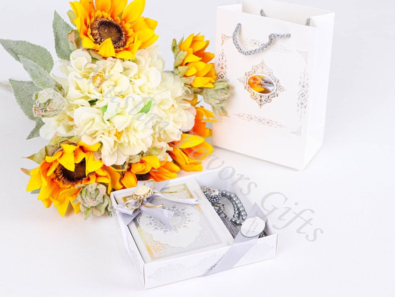 May include: A white gift box with a silver ribbon and a white gift bag. The box contains a small book and a silver prayer beads. A bouquet of sunflowers and white flowers is in the background.