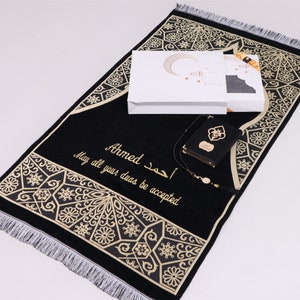 May include: A black prayer rug with gold embroidery and a white box with a crescent moon design. The rug has the name "Ahmed" and the text "May all your duas be accepted" embroidered on it. The set also includes a black prayer book and a prayer bead necklace.