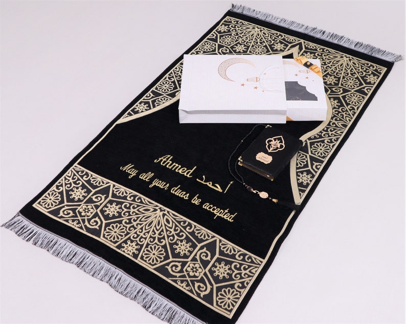 May include: A black prayer rug with gold embroidery and a white box with a crescent moon design. The rug has the name "Ahmed" and the text "May all your duas be accepted" embroidered on it. The set also includes a black prayer book and a prayer bead necklace.