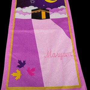 Peut inclure: Tapis de pri&egrave;re rectangulaire avec un motif violet et rose. Le haut repr&eacute;sente un ciel nocturne avec un croissant de lune, des &eacute;toiles et des nuages au-dessus d'un b&acirc;timent noir avec une porte dor&eacute;e. Le nom "Maryam" est brod&eacute; en rose.