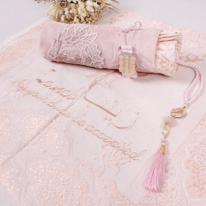May include: A pink prayer rug with a white fringe and a matching pink prayer mat rolled up in a pink pouch. The rug has a floral pattern and the words "In sha Allah, may your deeds be accepted" embroidered in gold thread. A pink prayer bead necklace with a gold charm hangs on the rug.