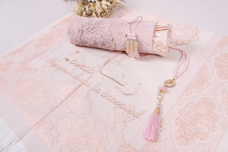 May include: A pink prayer rug with a white fringe and a matching pink prayer mat rolled up in a pink pouch. The rug has a floral pattern and the words "In sha Allah, may your deeds be accepted" embroidered in gold thread. A pink prayer bead necklace with a gold charm hangs on the rug.