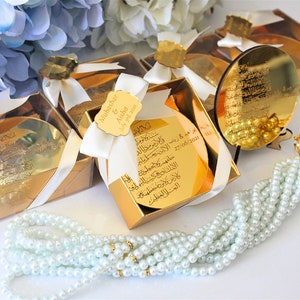 May include: Gold gift boxes with white ribbon and a small gold mirror with Arabic calligraphy. The boxes contain a small gold mirror with Arabic calligraphy. A white pearl prayer beads with a gold charm.