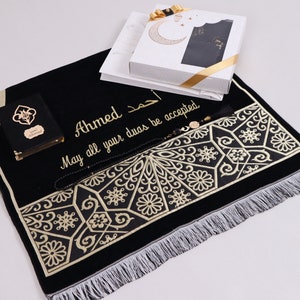 May include: A black prayer rug with gold embroidery and fringe. The rug has the name "Ahmed" and the text "May all your duas be accepted" embroidered on it. The rug is accompanied by a small black book, a black prayer bead necklace, and a white gift box.