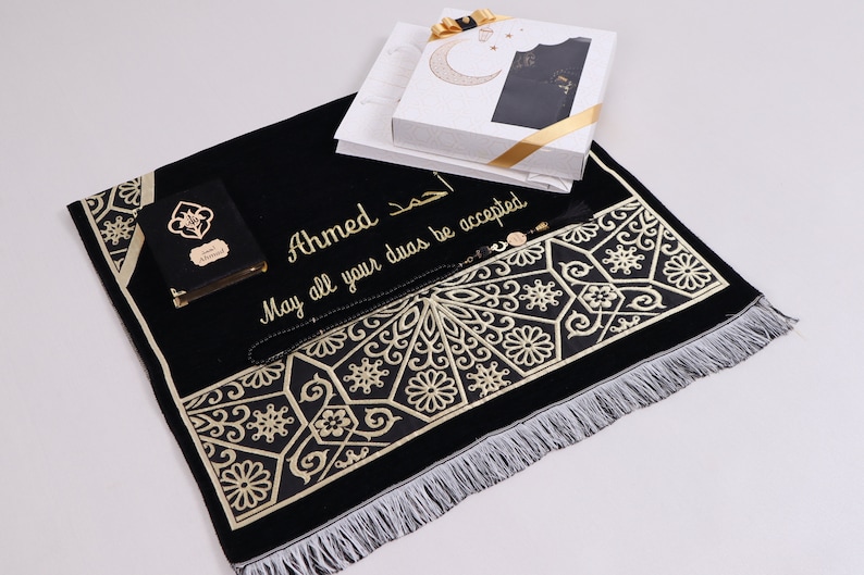 May include: A black prayer rug with gold embroidery and fringe. The rug has the name "Ahmed" and the text "May all your duas be accepted" embroidered on it. The rug is accompanied by a small black book, a black prayer bead necklace, and a white gift box.