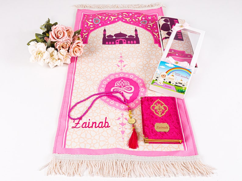 Peut inclure: Un tapis de pri&egrave;re rose avec un motif de mosqu&eacute;e, une bo&icirc;te assortie, un chapelet et un petit livre. Le tapis et le livre sont personnalis&eacute;s avec le nom "Zainab". Un bouquet de fleurs est &agrave; gauche.
