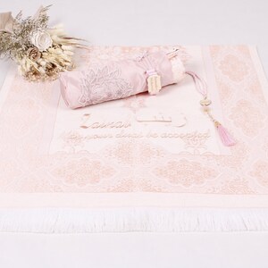 May include: A pink prayer rug with a white fringe, a matching pink prayer mat with a tassel, and the text "Insha'Allah May your deeds be accepted" embroidered on the rug.