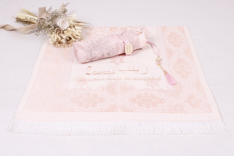 May include: A pink prayer rug with a white fringe, a matching pink prayer mat with a tassel, and the text "Insha'Allah May your deeds be accepted" embroidered on the rug.
