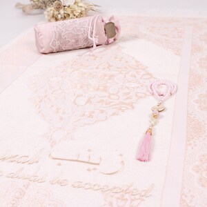 May include: A pink prayer rug with a floral design and the words "La ilaha illa Allah" in Arabic script. The rug is decorated with a pink tassel and a set of prayer beads.