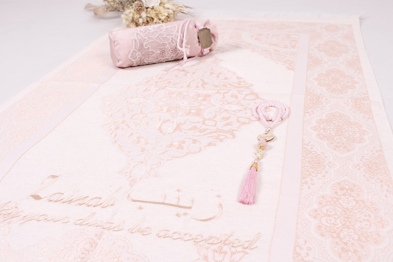 May include: A pink prayer rug with a floral design and the words "La ilaha illa Allah" in Arabic script. The rug is decorated with a pink tassel and a set of prayer beads.