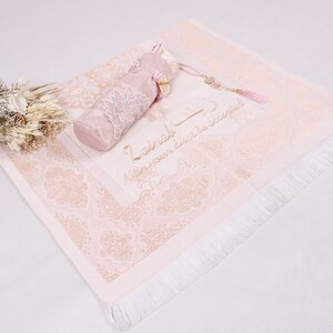 May include: A pink prayer rug with a white fringe and a matching pink prayer bead pouch. The rug has a floral pattern and the text "La ilaha illa Allah" which means "There is no god but Allah" in Arabic.