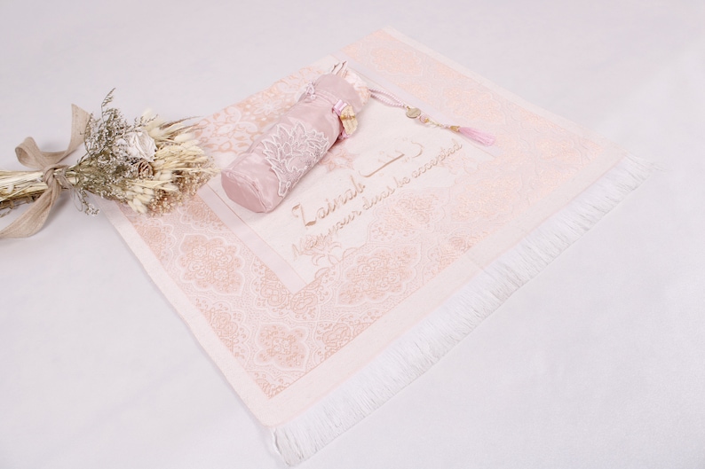 May include: A pink prayer rug with a white fringe and a matching pink prayer bead pouch. The rug has a floral pattern and the text "La ilaha illa Allah" which means "There is no god but Allah" in Arabic.