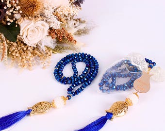 Könnte beinhalten: Zwei blaue Kristall-Gebetsperlen mit goldenen Quasten und Anhängern. Die Perlen befinden sich in einem weißen Spitzensäckchen mit einem goldenen Anhänger, auf dem "Masha Allah" steht.