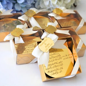 May include: Gold gift boxes with white ribbon bows and personalized gold mirror tags. The tags have Arabic calligraphy and the text "Mubashar & Aisha July grad. 2021".