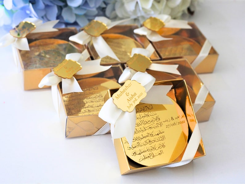May include: Gold gift boxes with white ribbon bows and personalized gold mirror tags. The tags have Arabic calligraphy and the text "Mubashar & Aisha July grad. 2021".