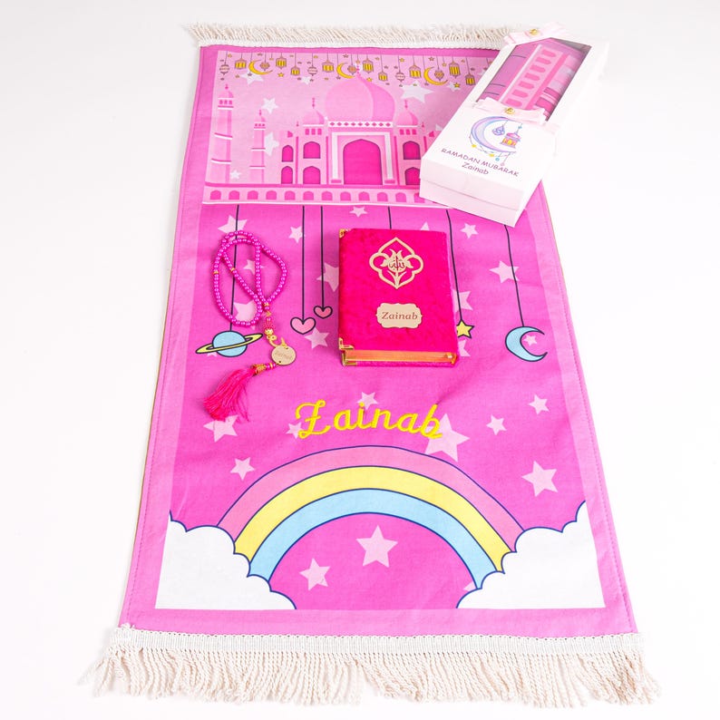May include: A pink prayer rug with a rainbow, stars, and a mosque design. Includes a pink Quran, prayer beads, and a gift box. The Quran is labeled "Zainab" in gold lettering. The rug has a white fringe.