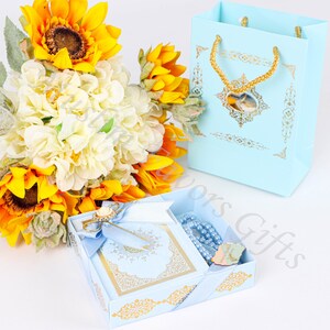 K&ouml;nnte beinhalten: Eine hellblaue Geschenkbox mit goldenen Akzenten enth&auml;lt ein kleines blaues Buch, eine blaue Gebetsperlenkette und eine goldene Nadel. Die Box ist von einem Strau&szlig; gelber Sonnenblumen und wei&szlig;er Hortensien umgeben.