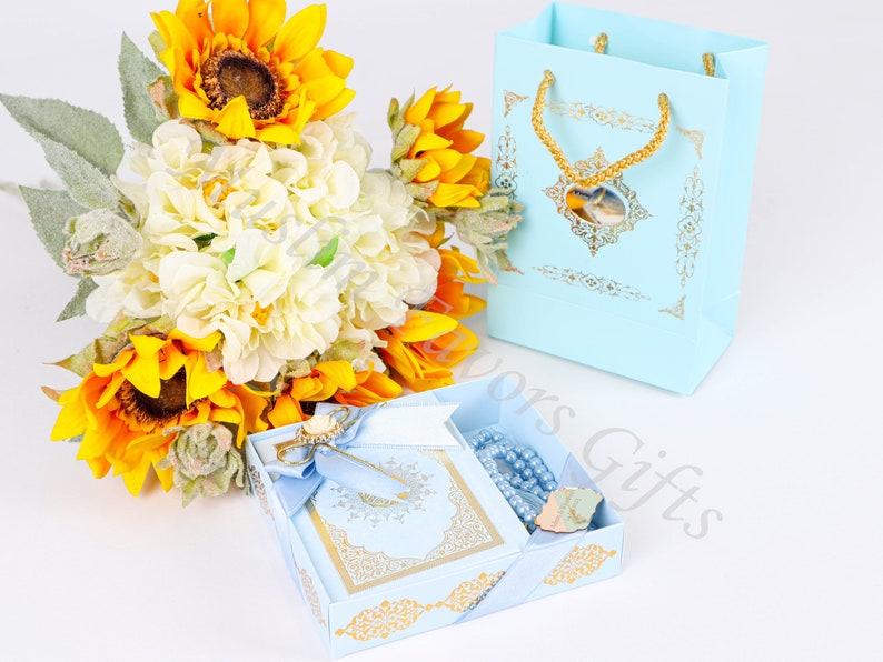 K&ouml;nnte beinhalten: Eine hellblaue Geschenkbox mit goldenen Akzenten enth&auml;lt ein kleines blaues Buch, eine blaue Gebetsperlenkette und eine goldene Nadel. Die Box ist von einem Strau&szlig; gelber Sonnenblumen und wei&szlig;er Hortensien umgeben.