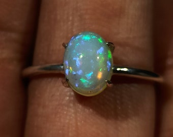 May include: A silver ring with an oval-shaped opal gemstone. The opal has a white base with iridescent green, blue, and purple flashes.