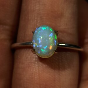 May include: A silver ring with an oval-shaped opal gemstone. The opal has a white base with iridescent green, blue, and purple flashes.