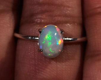 May include: A silver ring with an oval-shaped opal gemstone. The opal has a white base with flashes of green, red, and orange.
