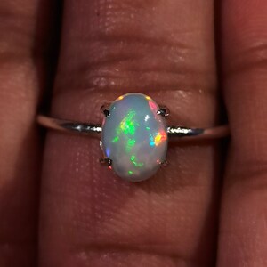 May include: A silver ring with an oval-shaped opal gemstone. The opal has a white base with flashes of green, red, and orange.