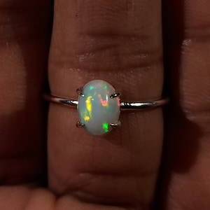 May include: A silver ring with an oval-shaped opal gemstone. The opal has a white base with flashes of green, yellow, and red.