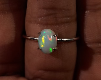 May include: A silver ring with an oval-shaped opal gemstone. The opal has a white base with flashes of green, yellow, and red.