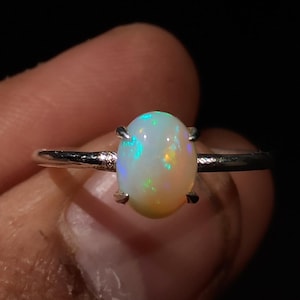 May include: A silver ring with an oval-shaped opal gemstone. The opal has a white base with iridescent flashes of green, blue, and yellow.
