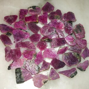 Può includere: Una collezione di pietre preziose di rubino grezze di varie forme e dimensioni. Le pietre grezze mostrano una gamma di tonalità rosa e rosso intenso, con alcuni accenni di verde e nero. Le pietre preziose sono sparse su una superficie bianca.